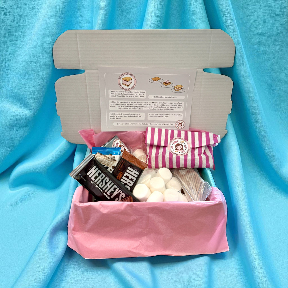 Open s’mores kit with burner, marshmallows and Hershey’s bars on blue fabric