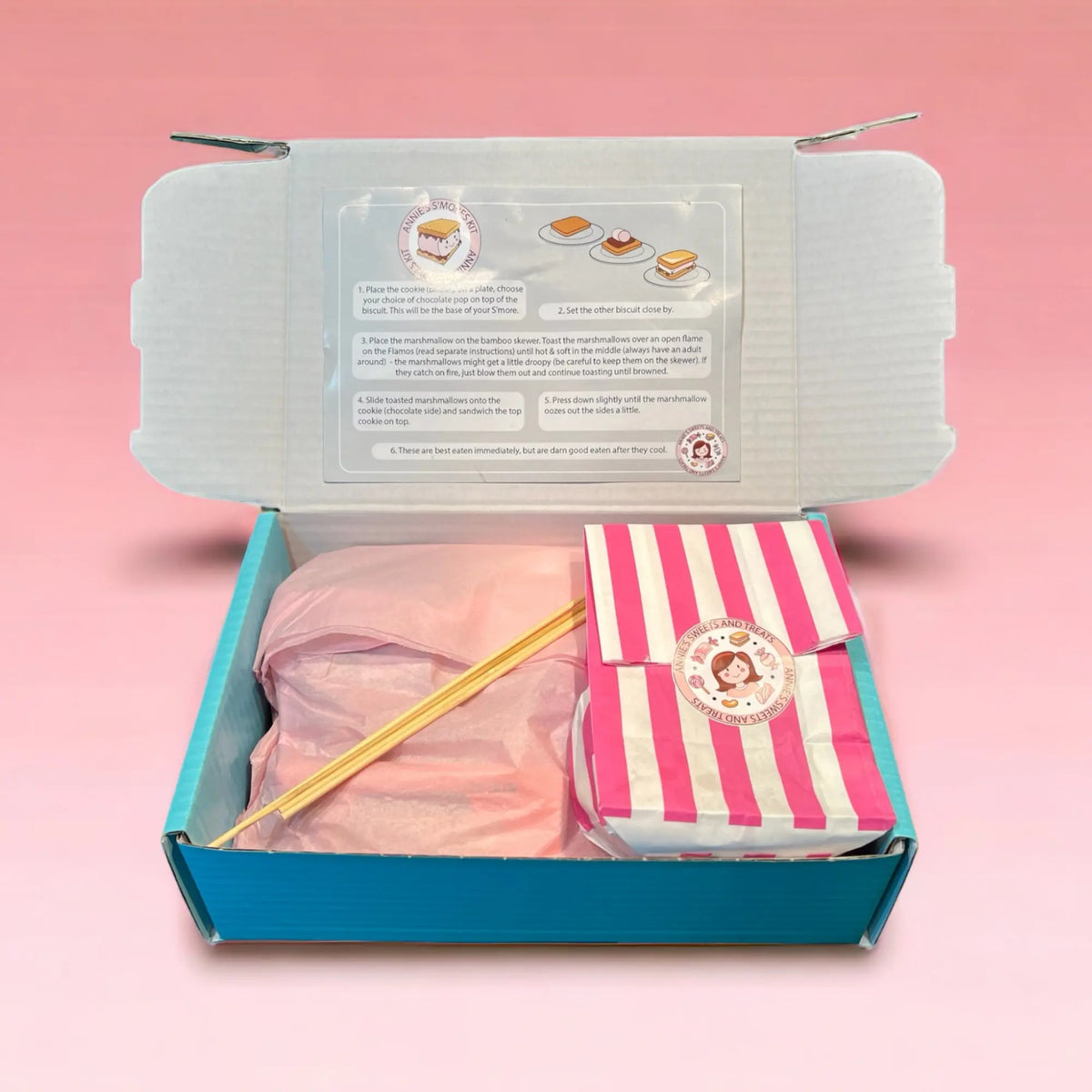Opened s’mores kit box showing the recipe card, skewers and pink candy-stripe bag
